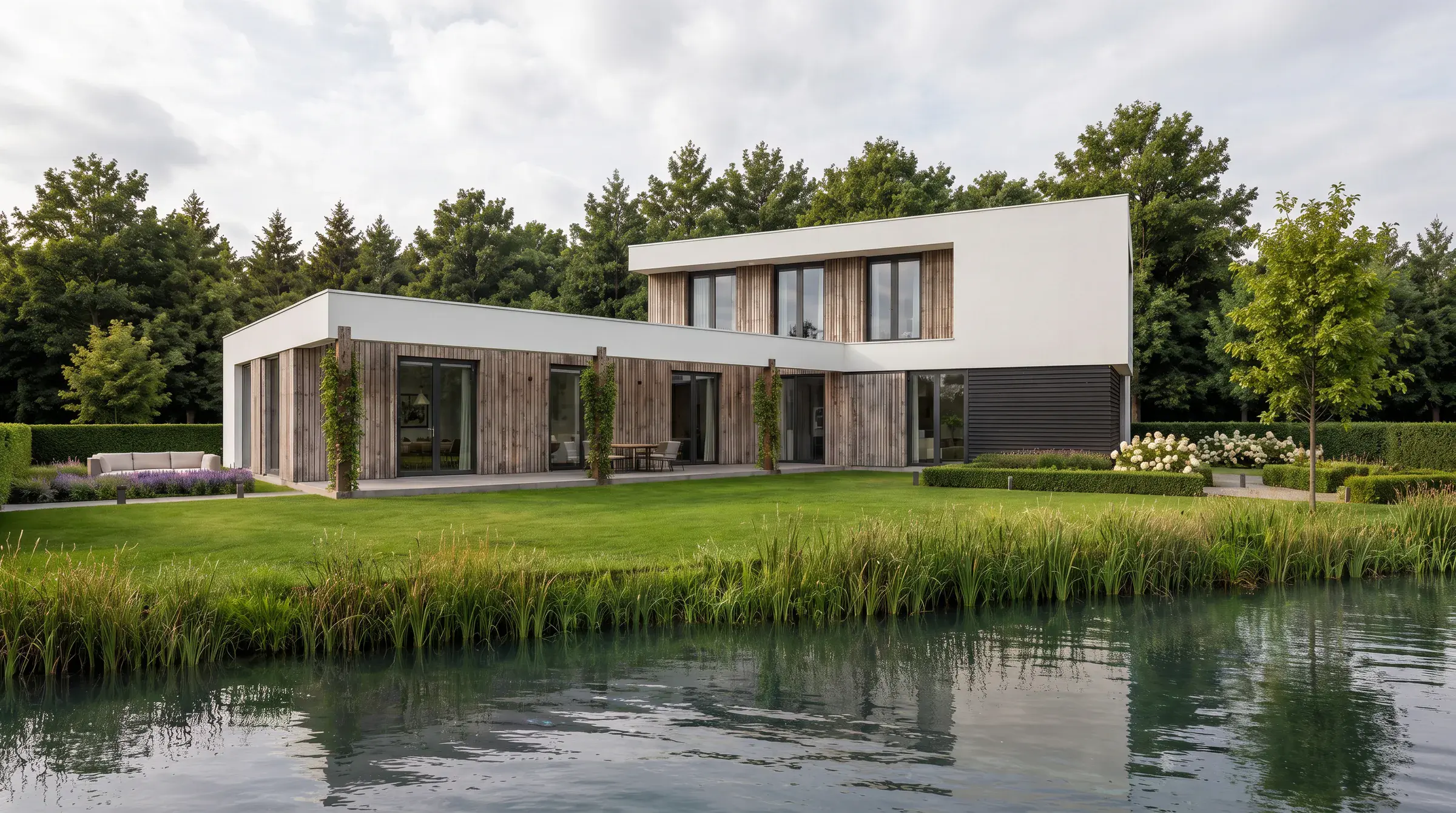 Impressie vanaf het water: een lage vleugel van hout en glas langs het gazon, daarboven een wit blok met hoge ramen en riet op de oever
